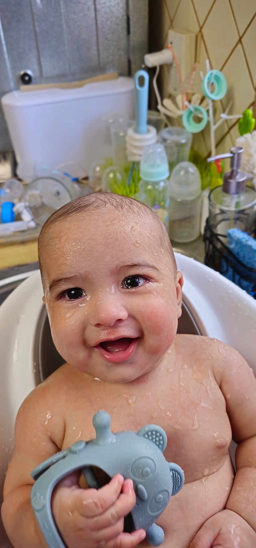 Adrian is registered to the contest to win money with this photo: baby, smiling, bath, water_droplets, toy, panda_toy, kitchen_sink, bottle, brush, wet_skin, child, happy, cute, indoors, person, closeup, face, hand, playful, cleaning
