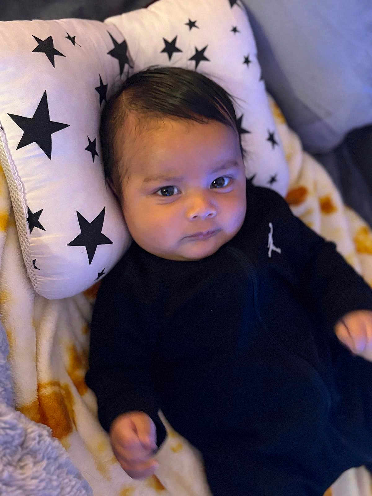 Zayan joined the competition — help win amazing prizes! baby, infant, pillow, blanket, black_clothing, face, head, person, cute, indoors, star_pattern, soft_fabric, child, young, portrait, lying_down, comfort, warm, expression, cozy