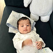 Olivia is registered to the contest to win money with this photo: baby, infant, onesie, stuffed_animal, couch, blanket, towel, diaper, indoor, child, person, cute, sleepwear, soft_toy, furniture, relaxing, lying_down, small_hands, face, calm