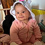 baby, child, pink_onesie, hood, smile, indoor, soft_toys, blanket, cozy, cute, face, infant, seated, wooden_furniture, pillow, play_area, warm_lighting, home, young_child, adorable