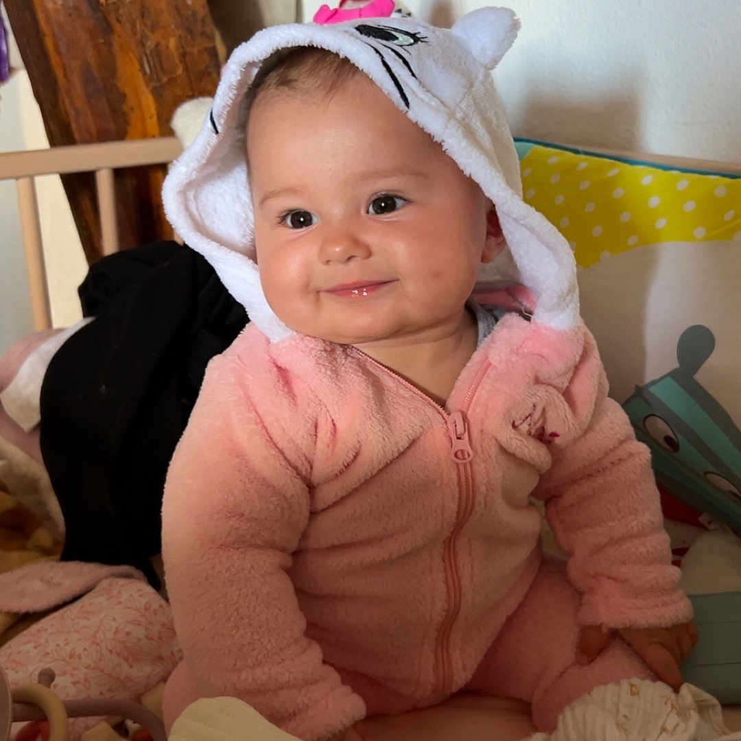 Maëlys Bodein a rejoint le concours — aidez-le/la à gagner de superbes lots ! adorable, baby, blanket, child, cozy, cute, face, home, hood, indoor, infant, pillow, pink_onesie, play_area, seated, smile, soft_toys, warm_lighting, wooden_furniture, young_child