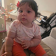 Jackeline joined the competition — help win amazing prizes! baby, child, infant, person, face, curly_hair, bow, earrings, orange_pants, pink_shirt, hand, indoor, furniture, couch, living_room, soft_light, cute, portrait, sitting, home