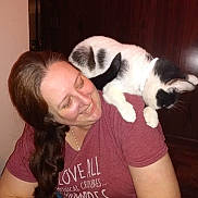 Toodles is registered to the contest to win money with this photo: woman, smiling, cat, black_and_white_cat, shoulder, indoor, wooden_wall, casual_clothing, long_hair, happy, pet, companion, portrait, human, animal, feline, domestic_animal, relaxed, friendship, person