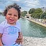 toddler, child, smiling, curly_hair, white_shirt, blue_shorts, river, water, stone_ledge, outdoor, cloudy_sky, trees, buildings, urban, hand_holding, happy, cute, portrait, daylight, person