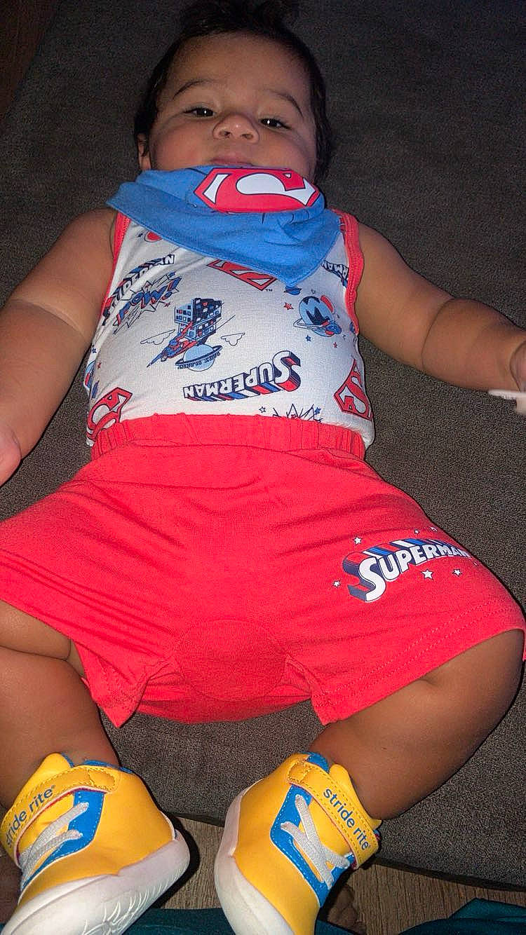 Kashton is registered to the contest to win money with this photo: baby, blue_scarf, carpet, child, clothing, costume, fashion, floor, indoor, infant, lying_down, photography, portrait, red_shorts, sitting, smiling, sneakers, superhero, superman, yellow_shoes