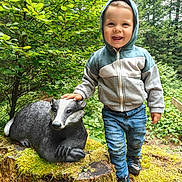 Léon a rejoint le concours — aidez-le/la à gagner de superbes lots ! child, toddler, hoodie, jeans, smiling, badger_statue, moss, tree_stump, forest, nature, outdoor, greenery, shoes, person, happy, daylight, casual_clothing, wood, foliage, cute
