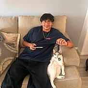 Mochi is registered to the contest to win money with this photo: beige, black_pants, casual, cat, cat_tag, chain_necklace, collar, couch, cushion, earrings, glasses, indoor, living_room, pet, phone, relaxed, smiling, tshirt, wood_floor, young_man