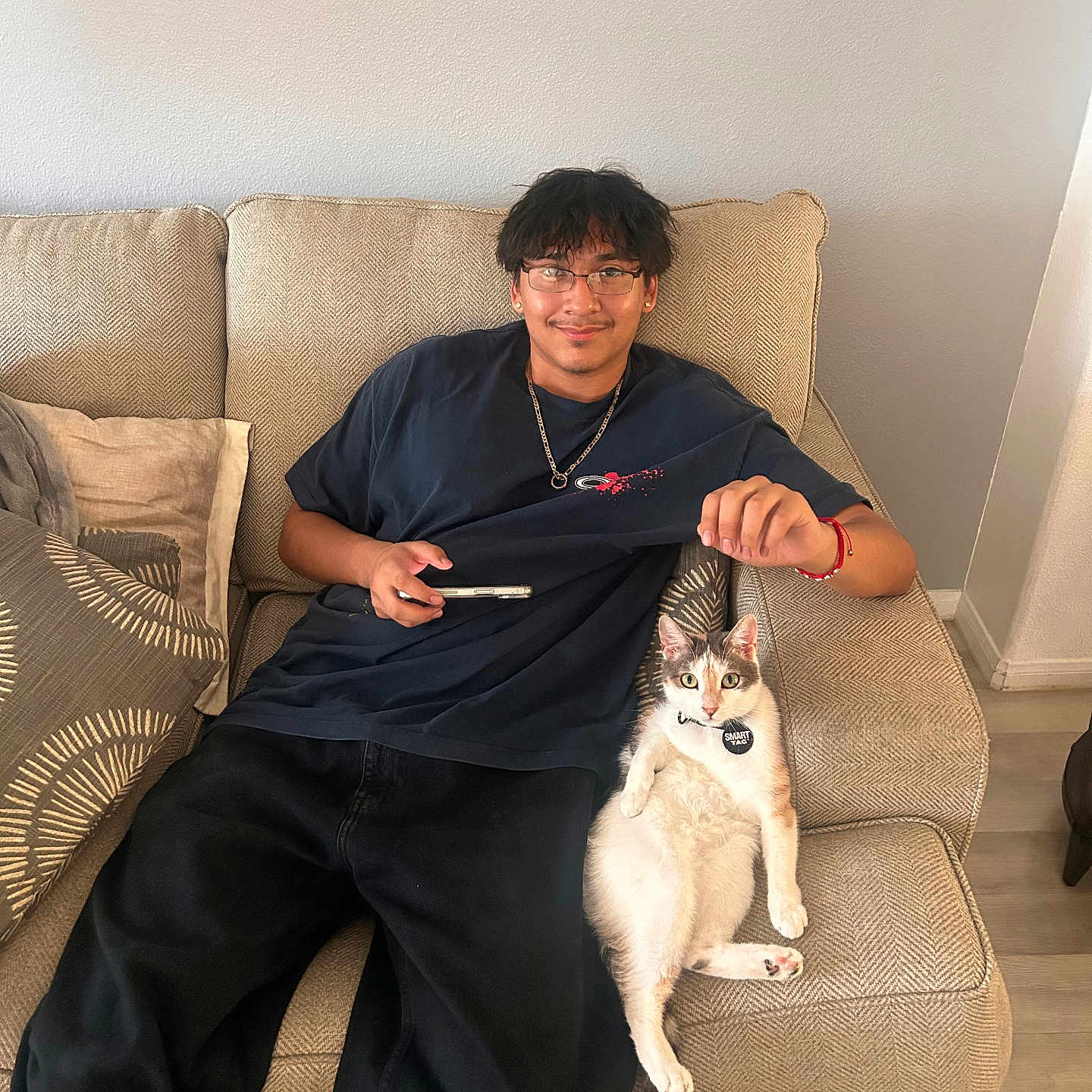 Mochi is registered to the contest to win money with this photo: beige, black_pants, casual, cat, cat_tag, chain_necklace, collar, couch, cushion, earrings, glasses, indoor, living_room, pet, phone, relaxed, smiling, tshirt, wood_floor, young_man