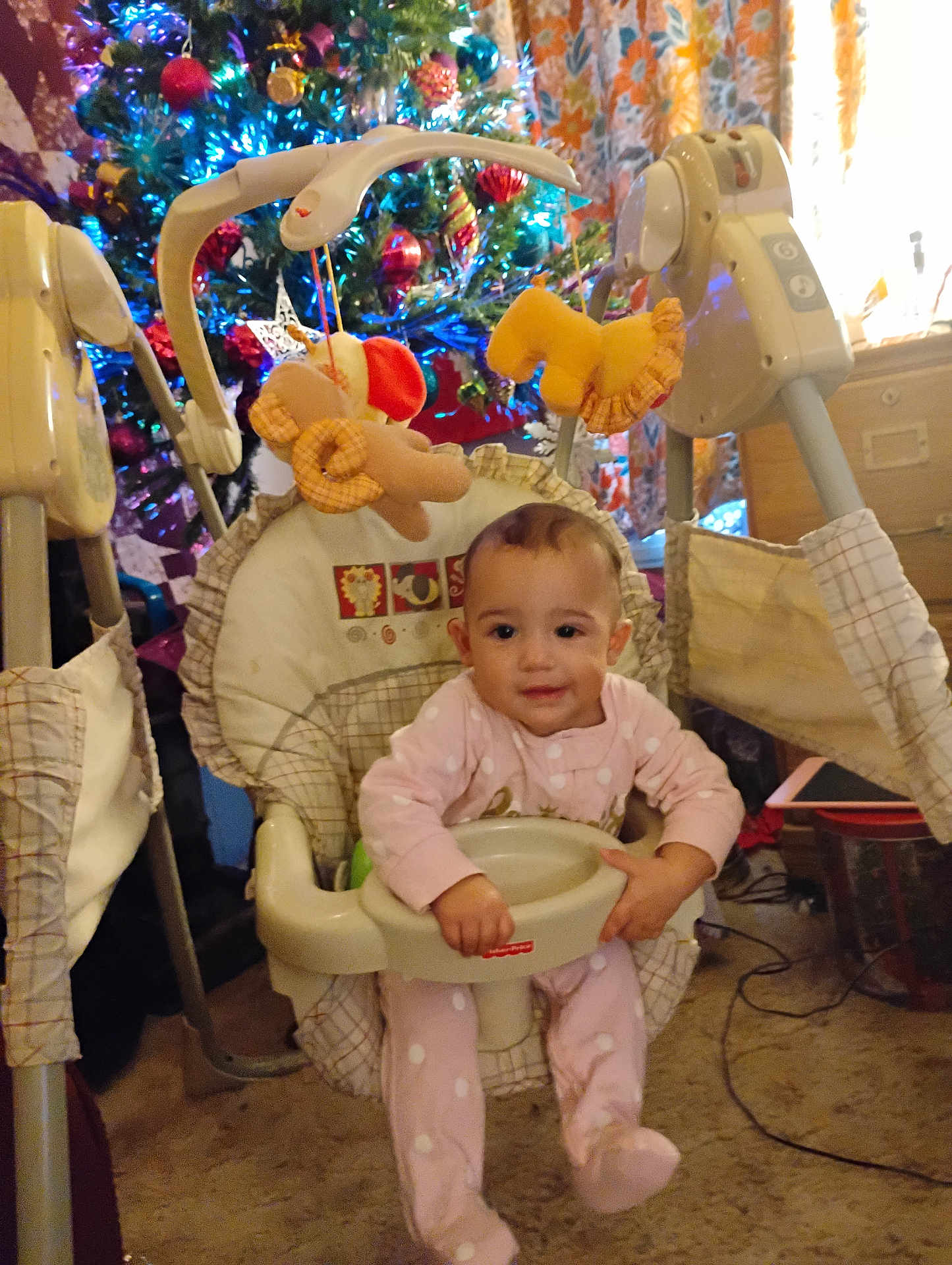 Athena is registered to the contest to win money with this photo: baby, child, infant, swing, baby_swing, christmas_tree, holiday_decor, lights, ornaments, plush_toy, indoor, carpet, furniture, curtains, smiling, pink_pajamas, face, person, festive, home