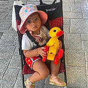Cassandre a rejoint le concours — aidez-le/la à gagner de superbes lots ! baby, child, cute, duck, footwear, hat, outdoor, pavement, person, playtime, red_shorts, sandals, sidewalk, sitting, stroller, summer_clothing, sunhat, toddler, toy, yellow_toy