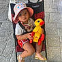 baby, child, cute, duck, footwear, hat, outdoor, pavement, person, playtime, red_shorts, sandals, sidewalk, sitting, stroller, summer_clothing, sunhat, toddler, toy, yellow_toy