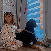 Arya participe au concours pour gagner de l'argent avec cette photo : child, dog, pajamas, indoor, floor, window, blinds, pet, calm, home, wooden_floor, black_dog, sitting, looking_out, hand_on_dog, cozy, relaxed, young_child, casual_clothing, domestic_scene
