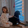 child, dog, pajamas, indoor, floor, window, blinds, pet, calm, home, wooden_floor, black_dog, sitting, looking_out, hand_on_dog, cozy, relaxed, young_child, casual_clothing, domestic_scene