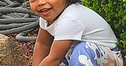 Na'Son is registered to the contest to win money with this photo: child, crouching, curly_hair, dark_hair, face, garden, happy, nature, outdoor, patterned_pants, plants, playful, portrait, rocks, shrubbery, sitting, smiling, sneakers, toddler, white_shirt