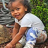 child, crouching, curly_hair, dark_hair, face, garden, happy, nature, outdoor, patterned_pants, plants, playful, portrait, rocks, shrubbery, sitting, smiling, sneakers, toddler, white_shirt