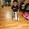 adult, black_hat, black_shirt, child, clothing, couch, family, furniture, gray_pants, indoor, living_room, nike_logo, phone, playtime, portrait, ride_on_toy, smiling, toddler, toy_vehicle, wooden_floor