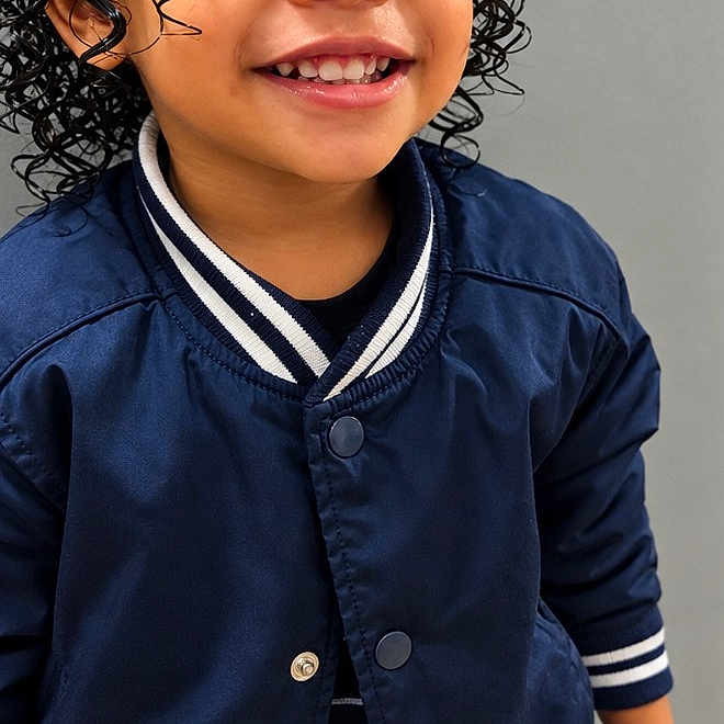 Adonis is registered to the contest to win money with this photo: child, toddler, smiling, curly_hair, portrait, indoors, jacket, navy_blue, varsity_jacket, striped_collar, striped_cuffs, beige_pants, standing, happy, face, eyes, teeth, fashion, close_up, photography