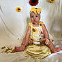 baby, child, cake, messy, frosting, sunflower, headband, dress, sitting, indoor, white_background, food, celebration, cute, portrait, hands, feet, yellow_bow, flower_decor, smudged
