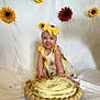 baby, child, cake, cake_smash, yellow_bow, sunflower, headband, dress, smiling, blue_eyes, white_backdrop, decorative_flowers, wooden_platter, messy, celebration, sitting, cute, infant, playful, portrait