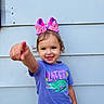 child, toddler, smiling, pointing, bow, pink, purple_shirt, crocodile_graphic, casual_clothing, outdoor, wall, wood_panel, happy, cute, portrait, person, short_hair, summer, playful, fashion
