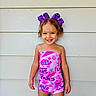 child, girl, smile, purple_bows, curly_hair, pink_outfit, patterned_clothing, standing, happy, portrait, wall, outdoor, casual, cute, fashion, person, toddler, bright, colorful, young