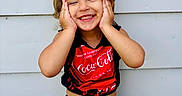 Mazikeen is registered to the contest to win money with this photo: toddler, child, smiling, red_bow, coca_cola, checkered_shorts, happy, portrait, outdoor, standing, hands_on_face, cute, fashion, headband, wall, stone, background, person, apparel, expression