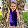child, girl, purple_outfit, bow, smiling, tree, grass, outdoor, happy, standing, nature, sunlight, playful, youth, feet, shoes, portrait, casual_clothing, daytime, cute