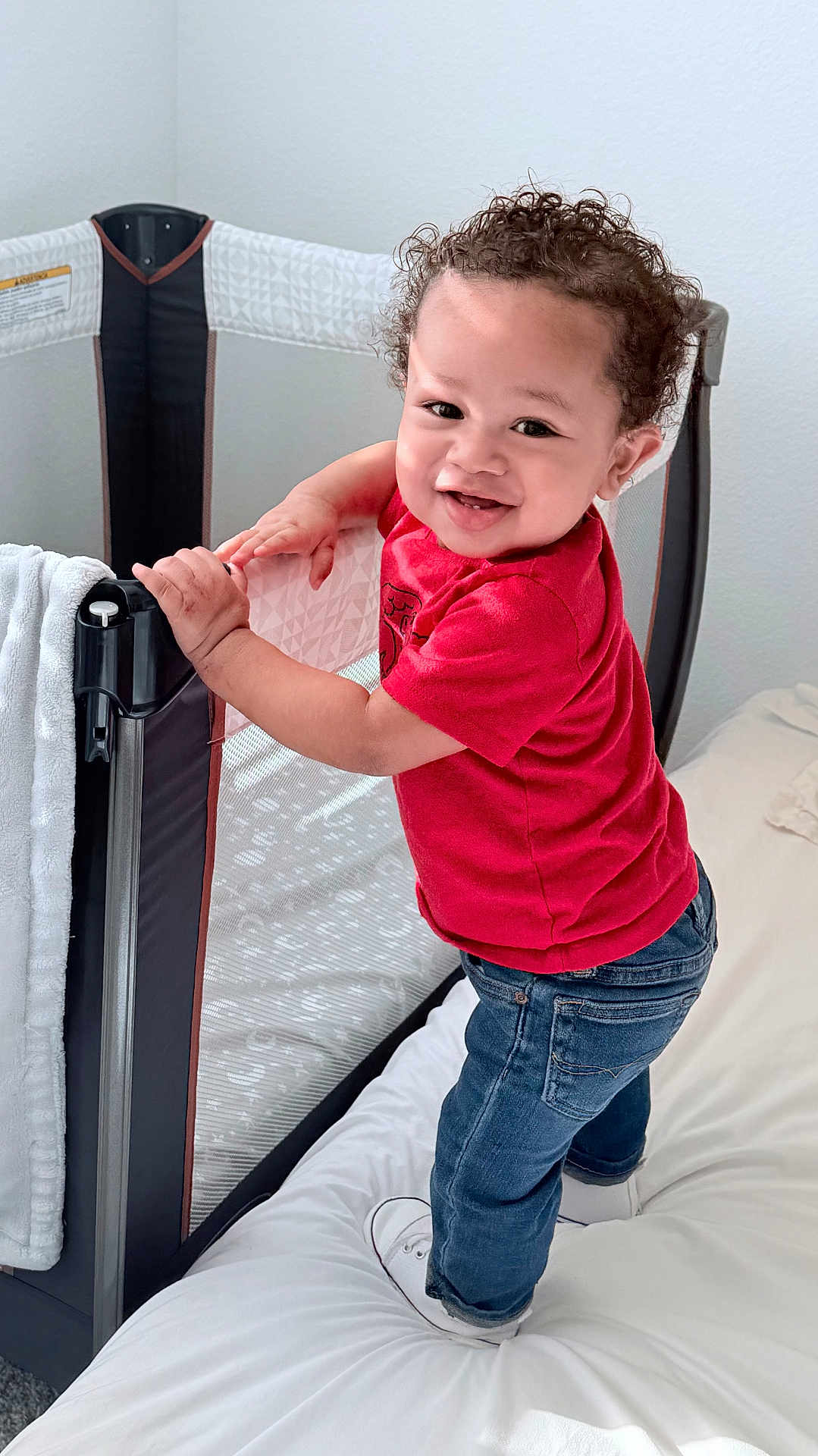 Elijah is registered to the contest to win money with this photo: toddler, child, smiling, red_shirt, jeans, curly_hair, bed, playpen, white_bedspread, indoor, happy, standing, person, baby, handrail, cot, footwear, casual_clothing, home, daylight