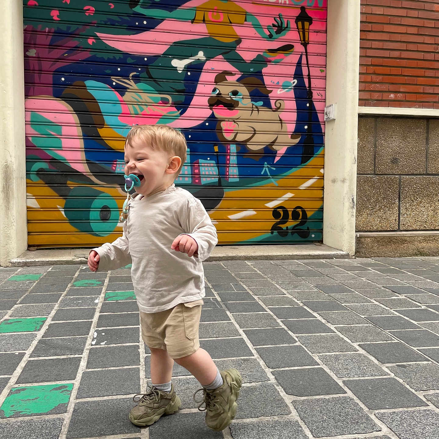 Amaë a rejoint le concours — aidez-le/la à gagner de superbes lots ! building, cartoon, child, cobblestone, dog, door, graffiti, long_sleeve_shirt, mural, outdoor, pacifier, playful, shorts, smiling, sneakers, street, toddler, urban_art, walking, wall