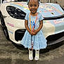 child, girl, blue_dress, sparkly_boots, smiling, standing, sports_car, white_car, cartoon_decorations, indoors, convention_center, lanyard, hair_bows, cute, happy, floor, license_tag, head, face, event