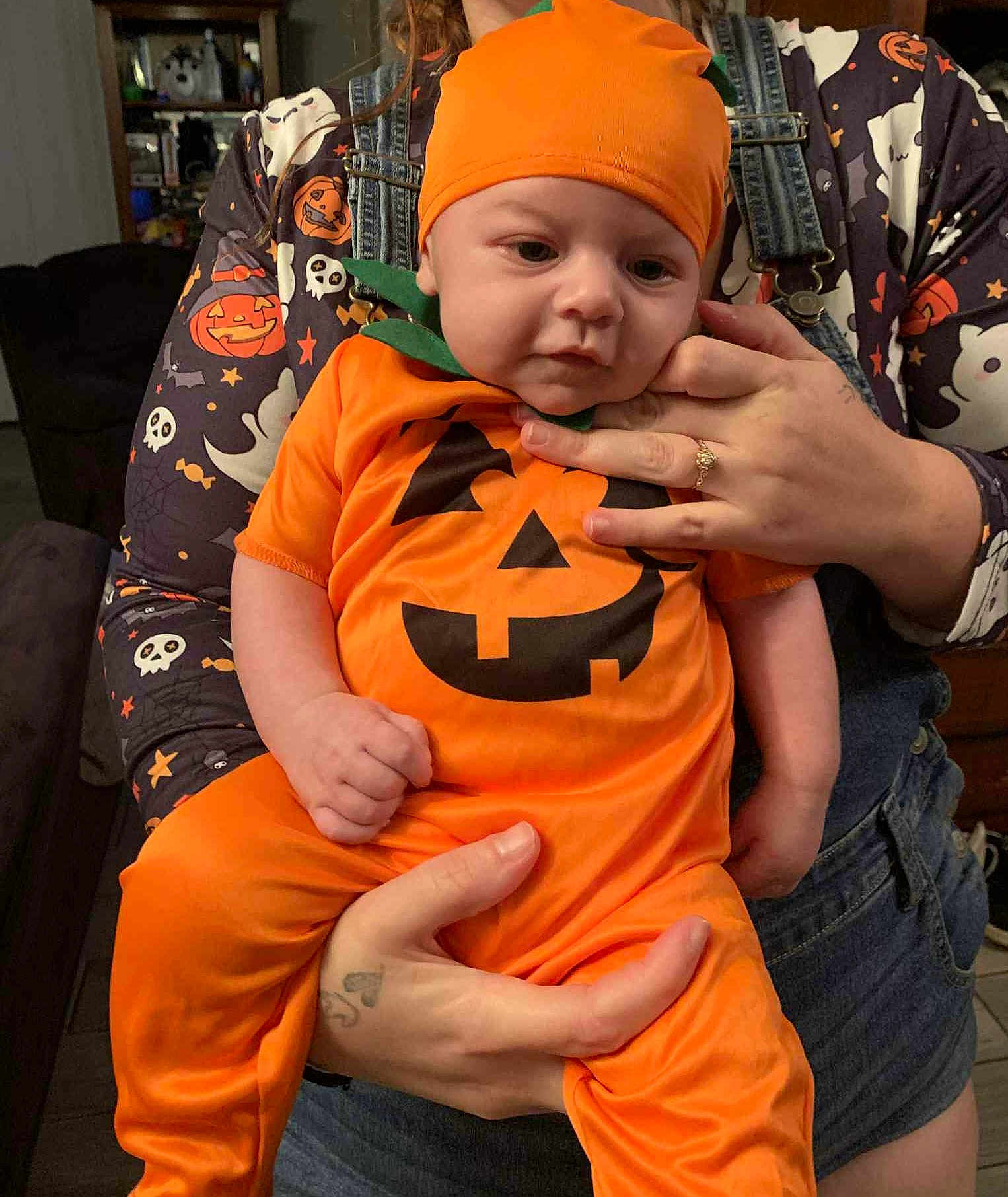 Grayson joined the competition — help win amazing prizes! baby, infant, pumpkin_costume, orange_clothing, halloween, child, costume, adult_hands, denim_overalls, tattoo, indoor, portrait, cute, festive, holding, calm, holiday, face, person, sitting