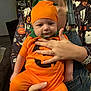 baby, pumpkin_costume, orange_clothing, halloween, infant, child, adult, holding, portrait, indoor, casual, cute, costume, festive, smile, face, person, hand, overalls, fun