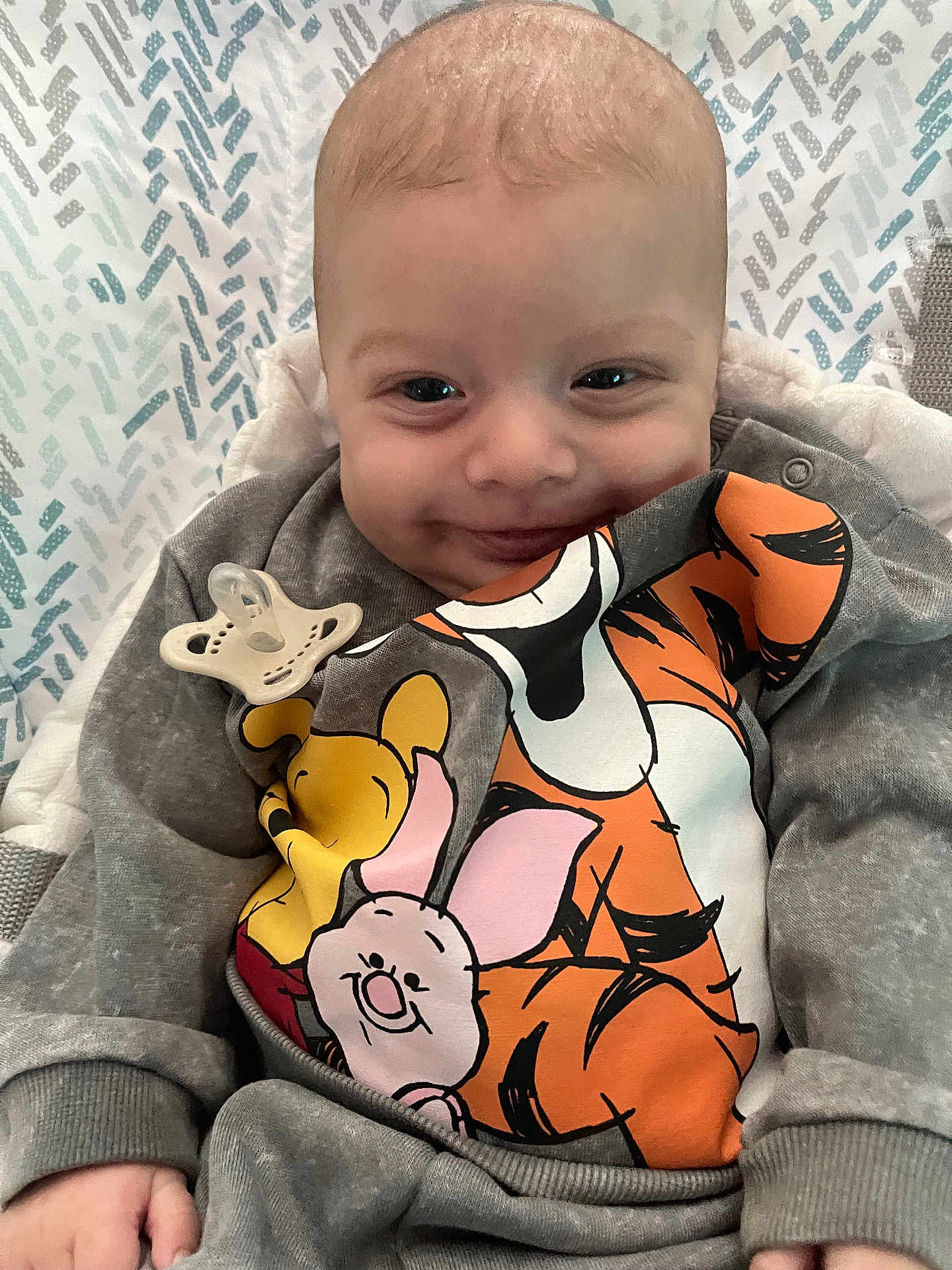 Grayson is registered to the contest to win money with this photo: baby, smiling, pacifier, grey_clothing, winnie_the_pooh, tigger, piglet, blanket, patterned_background, infant, cute, closeup, human, child, indoors, cozy, soft_lighting, portrait, happy, casual