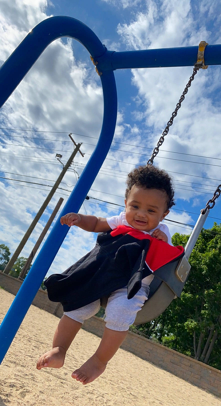 Travon is registered to the contest to win money with this photo: azure, blue, city, cloud, daytime, electric_blue, fun, happy, human_body, leisure, outdoor_play_equipment, people_in_nature, person, photograph, playground, recreation, sky, swing, toddler, tree