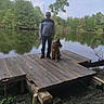 man, dog, german_shepherd, lake, dock, water, trees, nature, outdoor, sky, clouds, leash, wood, forest, reflection, casual_clothing, greenery, standing, pet, calm