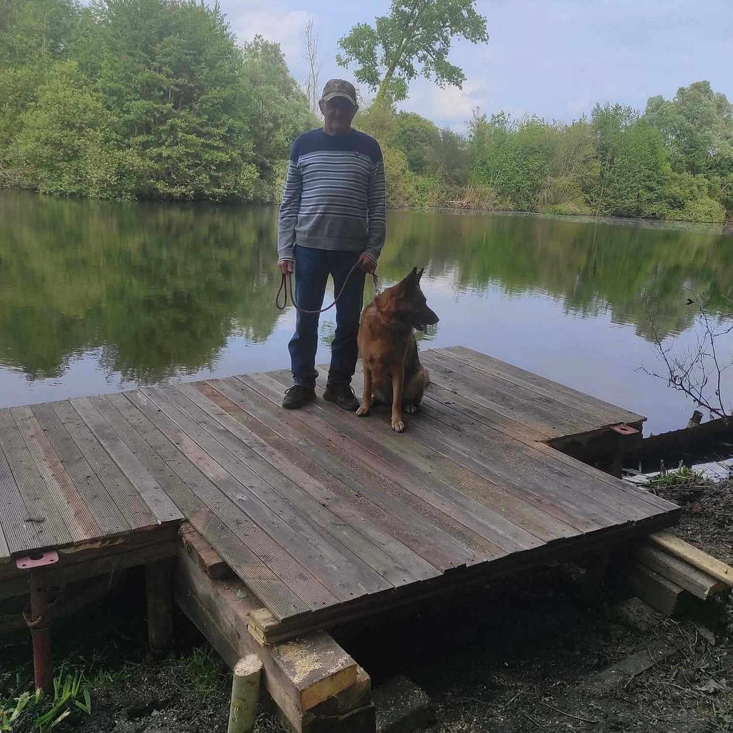 Alba participe au concours pour gagner de l'argent avec cette photo : man, dog, german_shepherd, lake, dock, water, trees, nature, outdoor, sky, clouds, leash, wood, forest, reflection, casual_clothing, greenery, standing, pet, calm