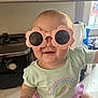Journey is registered to the contest to win money with this photo: baby, sunglasses, flower_shaped, pink, smiling, kitchen, counter, rainbow, green_shirt, purple_shorts, child, cute, playful, indoor, person, young_child, apparel, accessory, happy, portrait