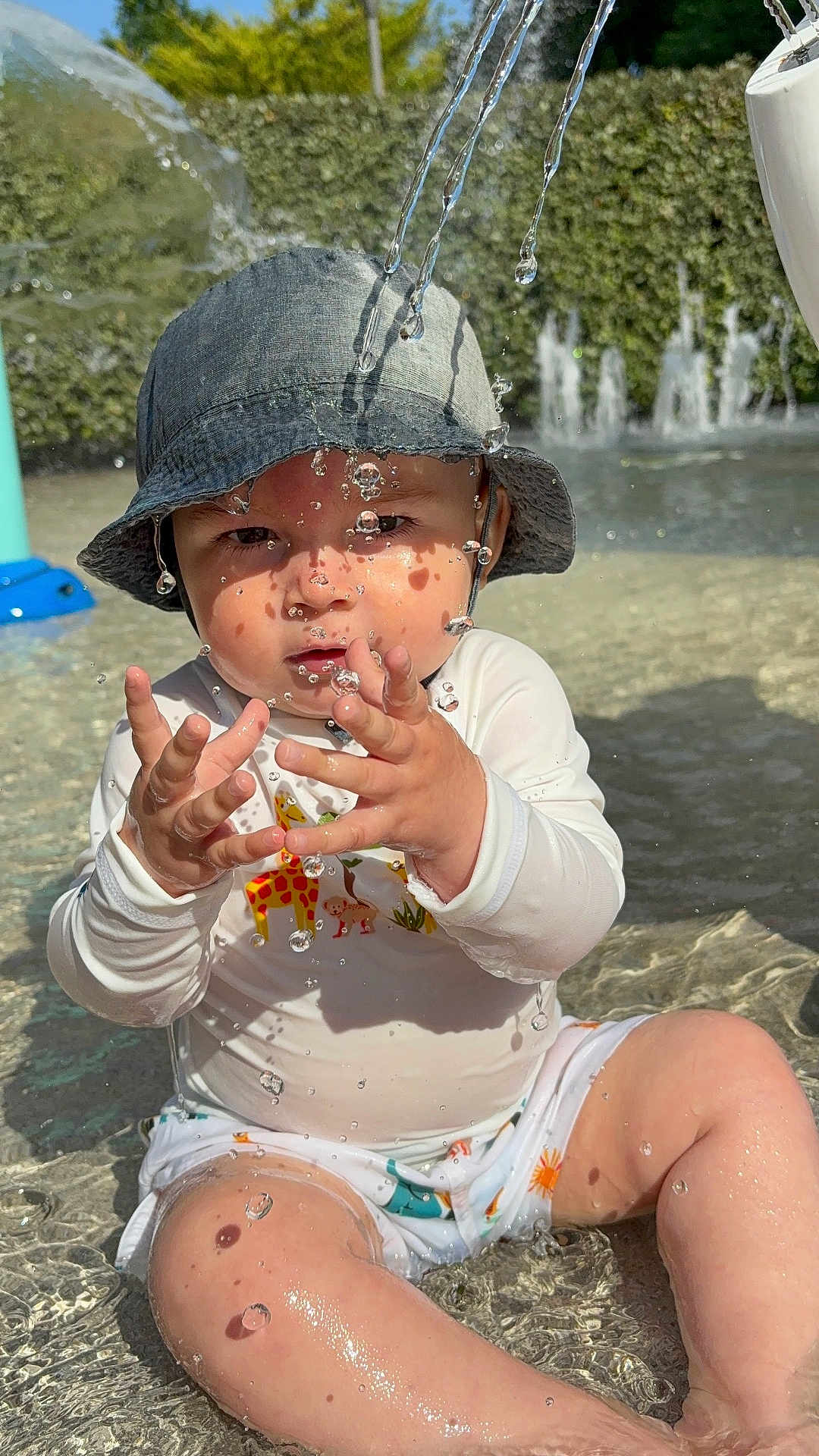 Andrea participe au concours pour gagner de l'argent avec cette photo : toddler, child, water, hat, bucket_hat, swimsuit, animal_print, playing, outdoor, pool, water_droplets, sunlight, reflection, summer, fun, sitting, cute, baby, face, hands
