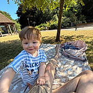 Oakley is registered to the contest to win money with this photo: child, toddler, mickey_mouse, shirt, blanket, picnic, park, tree, sunlight, grass, backpack, legs, person, outdoor, smiling, nature, bench, gazebo, shadow, reaching