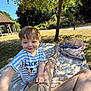 backpack, bench, blanket, child, gazebo, grass, legs, mickey_mouse, nature, outdoor, park, person, picnic, reaching, shadow, shirt, smiling, sunlight, toddler, tree