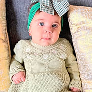 Amirah joined the competition — help win amazing prizes! baby, child, face, blue_eyes, headband, bow, green_dress, knitted_clothing, couch, pillow, indoor, portrait, cute, infant, sitting, small_hands, looking_at_camera, soft_lighting, cozy, expression