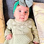 baby, child, face, blue_eyes, headband, bow, green_dress, knitted_clothing, couch, pillow, indoor, portrait, cute, infant, sitting, small_hands, looking_at_camera, soft_lighting, cozy, expression
