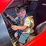 child, boy, cap, sunglasses, steering_wheel, race_car, red_car, seat, shorts, tshirt, backpack, smile, happy, vehicle_interior, seatbelt, daylight, playful, person, casual_clothing, indoor