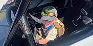 Hudson is registered to the contest to win money with this photo: child, cap, sunglasses, race_car, steering_wheel, seat, seatbelt, backpack, sunlight, shadow, outdoor, smile, person, vehicle, adventure, racing, helmet, metal, black, white
