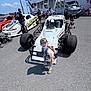 Hudson is registered to the contest to win money with this photo: child, race_car, racing, outdoor, asphalt, sunny, cap, sunglasses, stroller, adult, vehicle, race_track, people, car, toddler, medal, shorts, tshirt, shoes, backpack