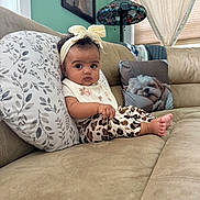 Athena is registered to the contest to win money with this photo: baby, child, couch, pillow, headband, leopard_print, indoors, living_room, cute, serious_expression, window, curtains, dog_pillow, green_wall, lamp, portrait, sitting, clothing, foot, decor