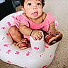 baby, child, curly_hair, pink_outfit, inflatable_seat, pink_bunnies, carpet, indoor, cute, tongue_out, shiny_shoes, white_cup, playful, seated, small_ears, bright_eyes, adorable, infant, hand_grip, portrait