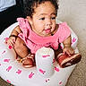 baby, bronze_shoes, bunny_pattern, carpet, child, curly_hair, cute, expression, face, furniture, indoor, infant, inflatable_seat, person, pink_dress, playful, room, shoes, sitting, small_child