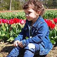 Ellie is registered to the contest to win money with this photo: child, toddler, denim_jacket, jeans, boots, curly_hair, tulips, flowers, garden, outdoor, spring, nature, plants, sunlight, portrait, sitting, greenery, daytime, cute, thoughtful