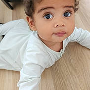 Eden a rejoint le concours — aidez-le/la à gagner de superbes lots ! baby, child, curly_hair, cute, expression, face, floor, hands, indoor, large_eyes, laying_down, long_sleeves, looking_up, person, portrait, skin, toddler, white_clothing, wooden_floor, young_child